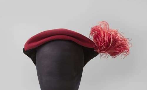 Hoed in een platte petvorm van rood laken en aan de zijkanten met zwart "gekreukeld" fluweel in twee driehoeken, aan één kant met speld met rode laken knop, rode veer by Hut Laco